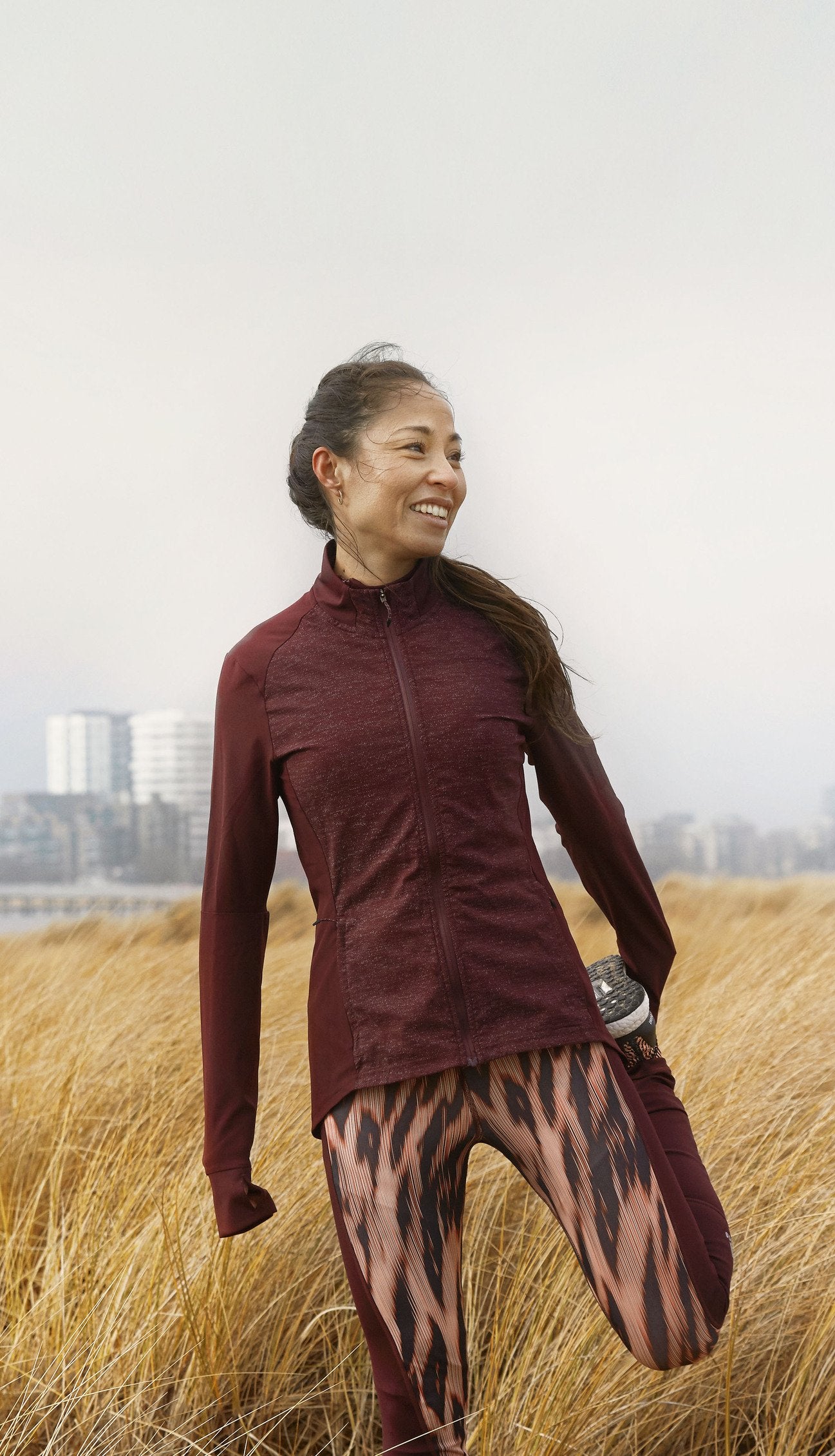 Smiling woman in a maroon zip-up jacket and patterned leggings jogging/stretching in a grassy field with a city skyline in the background.